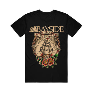 image of the front of a black tee shirt on a white background. tee has a full body print of a ship and flowers. across the top says bayside, just like home