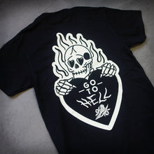 Load image into Gallery viewer, angled image of the back of a black tee shirt laid flat on a concrete floor. tee has a burning skeleton holding a heart that says go to hell