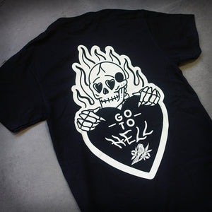 angled image of the back of a black tee shirt laid flat on a concrete floor. tee has a burning skeleton holding a heart that says go to hell