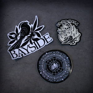 image of three stickers laid on a concrete floor. top sticker is a bird and says bayside. bottom sticker is a circle with a mandella and a bird design. the right sticker says go to hell and has a skeleton smoking a joint and the bird on it.