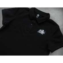 Load image into Gallery viewer, close up, angled image of a black polo shirtlaid flat on a concrete floor. front of the polo has a small white embroidered bird on the right chest 