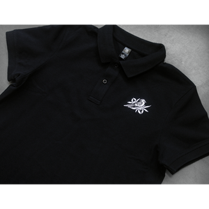 close up, angled image of a black polo shirtlaid flat on a concrete floor. front of the polo has a small white embroidered bird on the right chest 