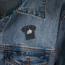 Load image into Gallery viewer, A Bayside enamel pin shaped like a T-shirt with a white bee design and secure rubber backing fastened above the chest pocket on the left side of a blue denim jacket.