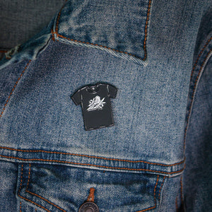 A Bayside enamel pin shaped like a T-shirt with a white bee design and secure rubber backing fastened above the chest pocket on the left side of a blue denim jacket.