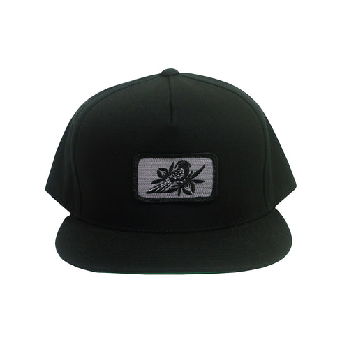 image of a black snapback hat on a white background. hat has small rectangle patch sewn on the front center. patch is grey with a black bird