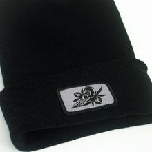 Load image into Gallery viewer, close up, angled image of a black winter beanie on a white background. beanie has sewn on rectangle patch on the front cuff. patch is grey with a bird in black