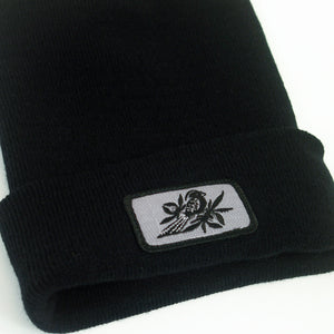 close up, angled image of a black winter beanie on a white background. beanie has sewn on rectangle patch on the front cuff. patch is grey with a bird in black