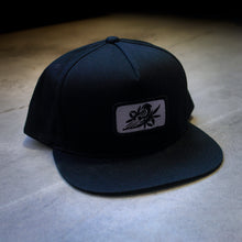 Load image into Gallery viewer, image of a black snapback hat on a concrete floor. hat has small rectangle patch sewn on the front center. patch is grey with a black bird