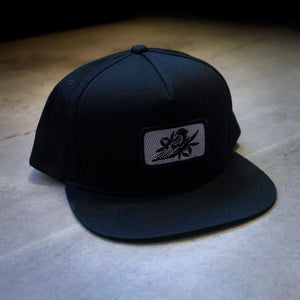 image of a black snapback hat on a concrete floor. hat has small rectangle patch sewn on the front center. patch is grey with a black bird