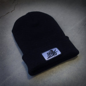 image of a black winter beanie laid flat on a concrete floor. beanie has sewn on rectangle patch on the front cuff. patch is grey with a bird in black