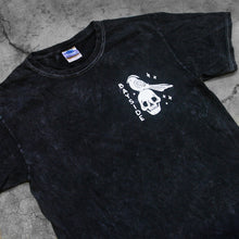 Load image into Gallery viewer, close up, angled image of the front of a black mineral wash tee shirt on a concete background. tee has a small chest print in white of a skull head and a bird. 