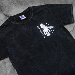 close up, angled image of the front of a black mineral wash tee shirt on a concete background. tee has a small chest print in white of a skull head and a bird. 