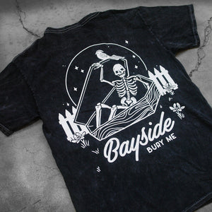 close up, angled image of the back of a black mineral wash tee shirt on a concrete background. tee has a back print in white of a skeleton opening up a coffin. below that says bayside bury me