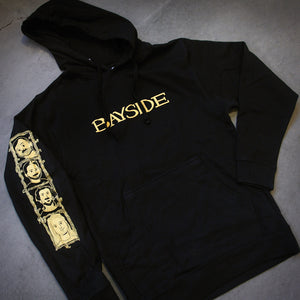 image of the front of a black pullover hoodie laid flat on a concrete floor. front of hoodie says bayside across the chest and has a left sleeve print. 