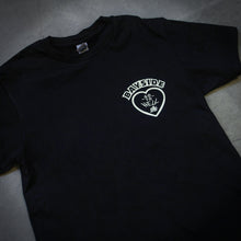 Load image into Gallery viewer, angled image of the front of a black tee shirt laid flat on a concrete floor. tee has a small right chest print of a heart and says bayside at the top