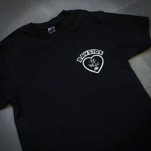 angled image of the front of a black tee shirt laid flat on a concrete floor. tee has a small right chest print of a heart and says bayside at the top