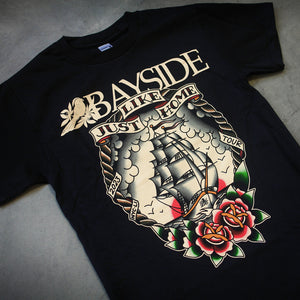 image of the front of a black tee shirt laid flat on a concrete floor. tee has a full body print of a ship and flowers. across the top says bayside, just like home