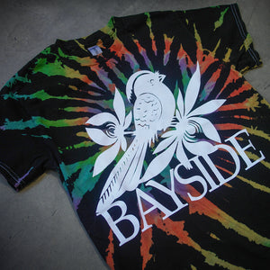 image of a black with rainbow spiral tie dye tee shirt laid flat on a concrete floor. front of the tee has a center print of a white bird that says bayside