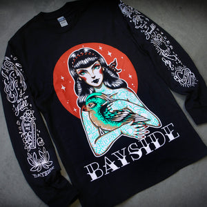 image of a black long sleeve tee shirt laid flat on a concrete floor. tee has full body print of a girl with tattoos all over holding a bird. across the bottom says Bayside. each sleeve has tattoo flash art