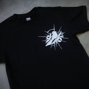 angled image of the front of a black tee shirt laid flatg on a concrete floor. tee has a small right chest print of a bird.