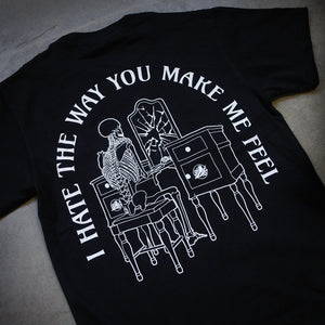 angled image of the back of a black tee shirt laid flat on a concrete. tee has a print of a person looking in a broken mirror of a vanity. arched around says i hate the way you make me feel.