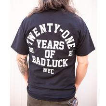 Load image into Gallery viewer, image of the back of a man in front of a white background. he has shoulder length hair draped in front of him and both hands in his olive green pants pockets. he has a tattoo on the left arm, and is wearing a black tee shirt that has a full back print in white. arch text that top says twenty one with the words years of bad luck N Y C stacked below. 