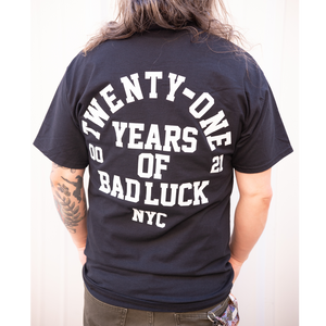 image of the back of a man in front of a white background. he has shoulder length hair draped in front of him and both hands in his olive green pants pockets. he has a tattoo on the left arm, and is wearing a black tee shirt that has a full back print in white. arch text that top says twenty one with the words years of bad luck N Y C stacked below. 