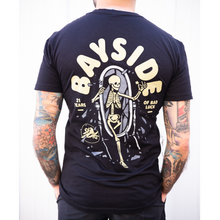 Load image into Gallery viewer, image of man from the neck down with tattoos on both arms and hands in pocket facing away showing the back of a black tee shirt. back of t shirt is on the left and has a full back print with grey skeleton stepping one leg out of a full length mirror. arched above in cream says bayside. on the left says 21 years and the right says of bad luck. there is a small circle and bird on the bottom left of the design.