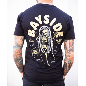 image of man from the neck down with tattoos on both arms and hands in pocket facing away showing the back of a black tee shirt. back of t shirt is on the left and has a full back print with grey skeleton stepping one leg out of a full length mirror. arched above in cream says bayside. on the left says 21 years and the right says of bad luck. there is a small circle and bird on the bottom left of the design.