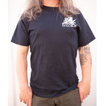 Load image into Gallery viewer, image of a man standing in front of a white background from the neck down. he has shoulder length brown hair, and olive green pants. he has a tattoo on the right arm and thumb in pocket. wearing a black tee shirt with a right chest print of a bird on leaves over the word bayside in white print.