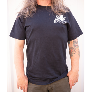 image of a man standing in front of a white background from the neck down. he has shoulder length brown hair, and olive green pants. he has a tattoo on the right arm and thumb in pocket. wearing a black tee shirt with a right chest print of a bird on leaves over the word bayside in white print.