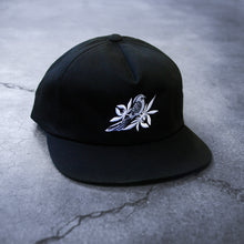 Load image into Gallery viewer, image of a black snapback on a concrete background. hat has white embroidery of a bird with leaves