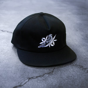image of a black snapback on a concrete background. hat has white embroidery of a bird with leaves