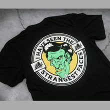 Load image into Gallery viewer, close up, angled image of the back of a black tee shirt on a concrete background. tee has a print of a green, zombie face. around the face says i have seen the strangest faces