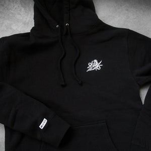 image of a black pullover hoodie laid flat on a concrete floor. small white embroidered bird on the right chest and a small sewn on patch that says bayside on the left sleeve