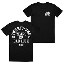 Load image into Gallery viewer, image of the front and back of a black tee shirt on a white background. the back of the shirt is on the left and has a full back print in white. arch text that top says twenty one with the words years of bad luck N Y C stacked below. the front of the shirt is on the right and has a small chest print on the right of a bird and leaves on top of the word bayside in white print.