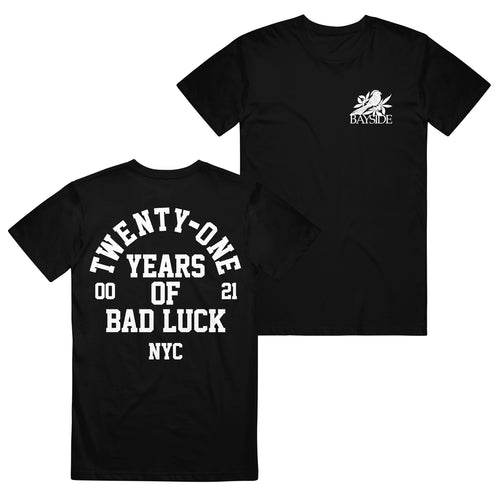 image of the front and back of a black tee shirt on a white background. the back of the shirt is on the left and has a full back print in white. arch text that top says twenty one with the words years of bad luck N Y C stacked below. the front of the shirt is on the right and has a small chest print on the right of a bird and leaves on top of the word bayside in white print.
